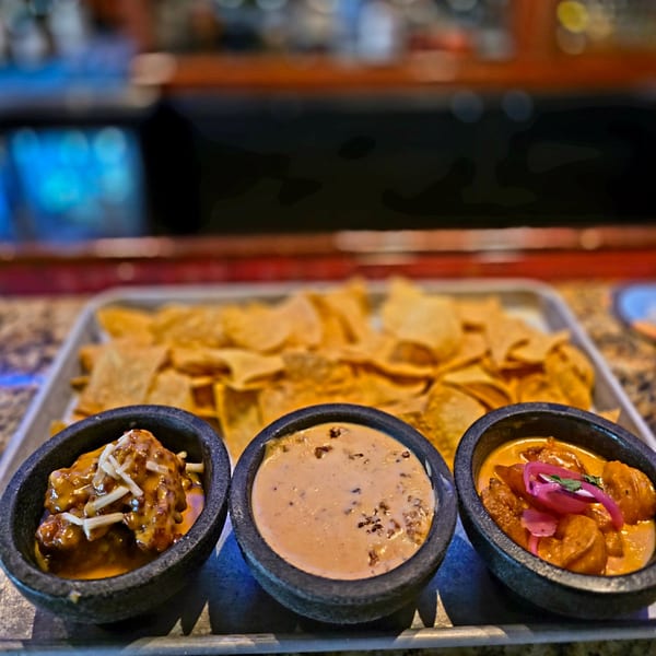 Three bowls of dips and sauces, including queso, with a tray of tortilla chips in the background.
