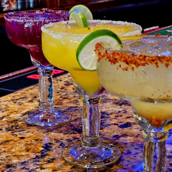 Three colorful margaritas, one purple, one yellow, and one light yellow with chili powder, on a bar.
